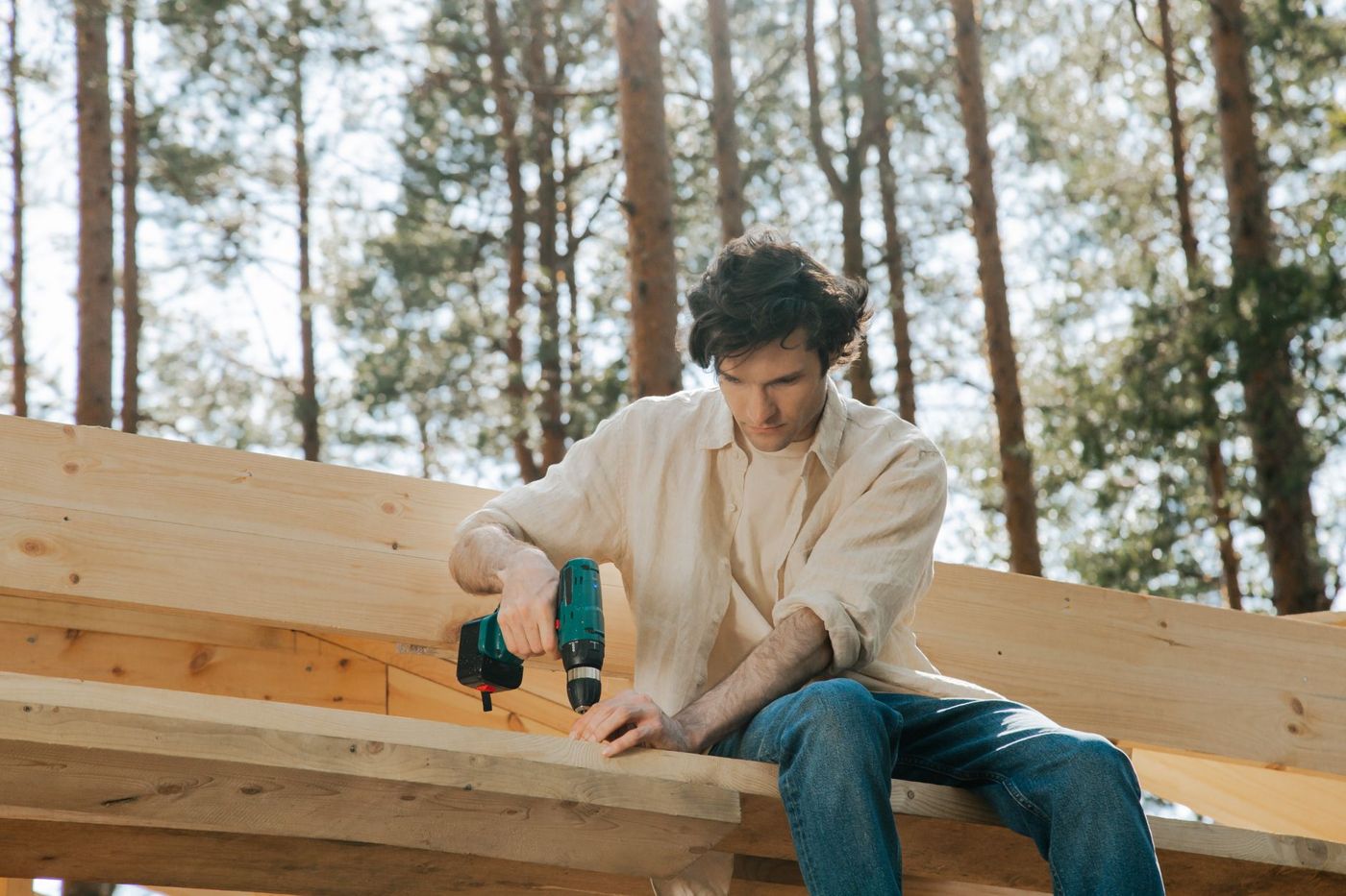 A carpenter in a linen shirt driving a screw into framing lumber with a cordless drill, tall pines behind him