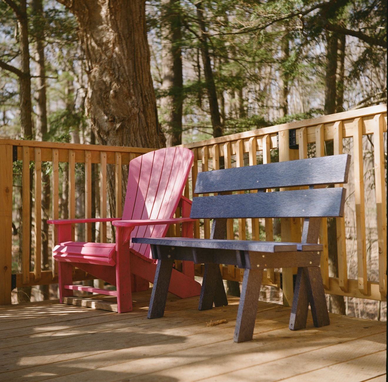 A pink Adirondack chair beside a dark slatted bench on a pale wood deck with a vertical-baluster railing, forest behind