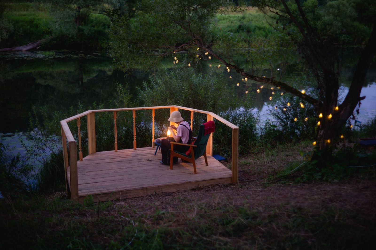 A small wooden deck with a railing beside a river at dusk, a person reading by lantern light under string lights in a tree