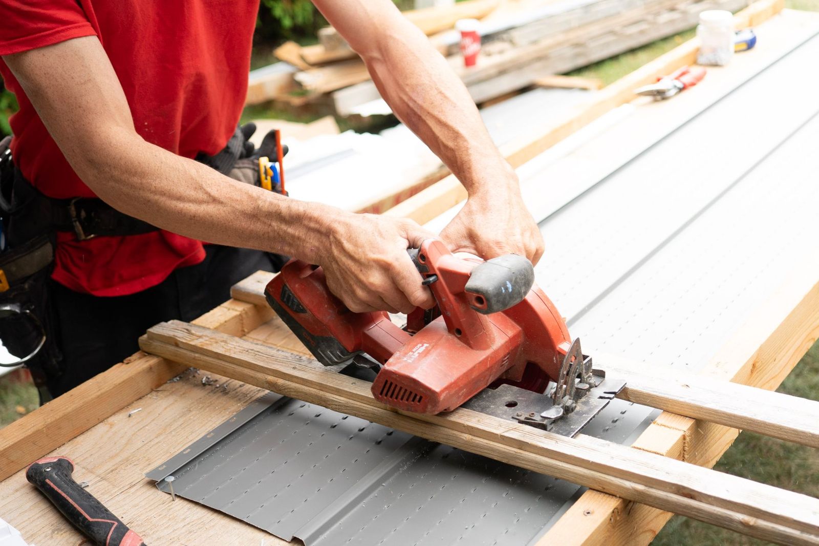 Close-up of a carpenter in a red shirt cutting a board with a cordless circular saw on a worktable outdoors