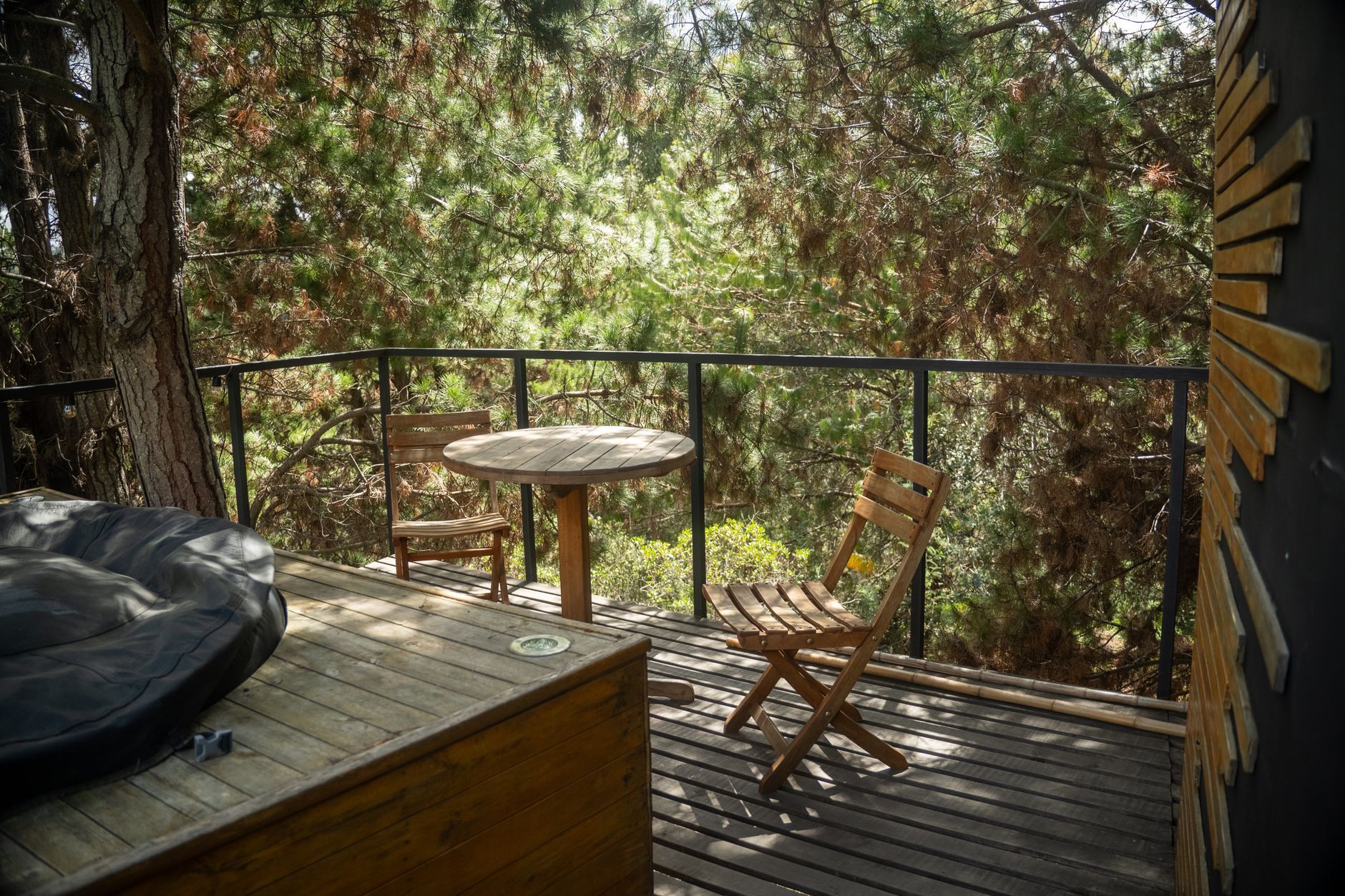A dark-stained deck tucked among pine trees, with a round wooden table, two folding chairs and a slim black metal railing