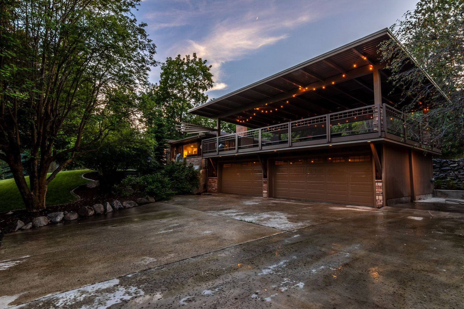 A covered upper deck with string lights and a dark metal-framed railing above a two-car garage at dusk, trees all around