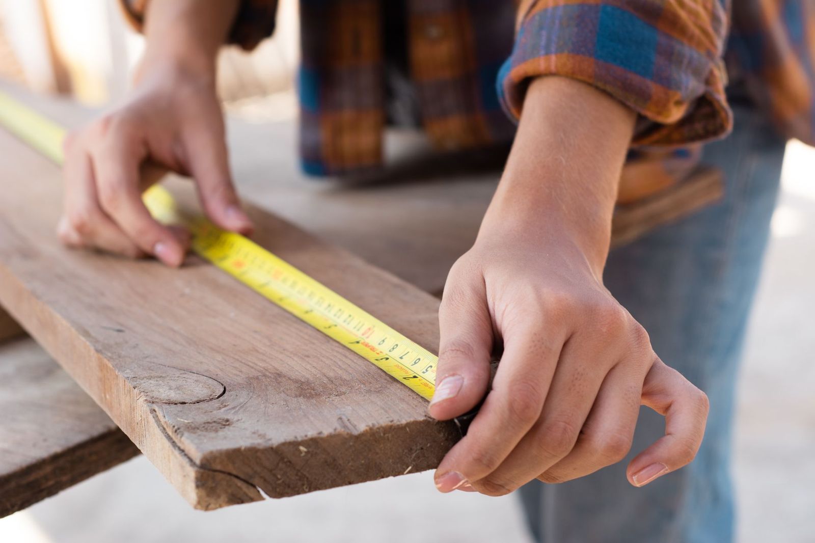 Close-up of hands holding a yellow tape measure along the edge of a weathered wooden plank