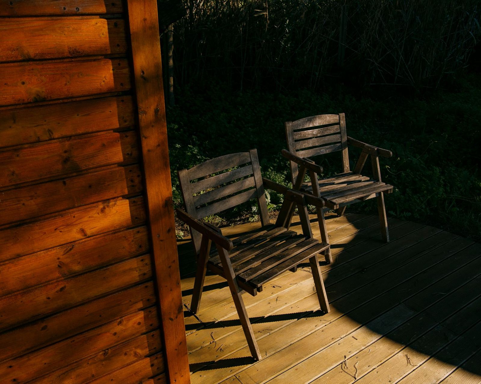 Two weathered wooden armchairs on a light wood deck beside a cabin wall of warm orange boards, lit by low sun